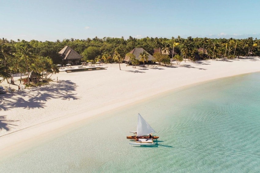 A sailboat glides over clear turquoise water near a tropical beach with palm trees and huts under a blue sky.