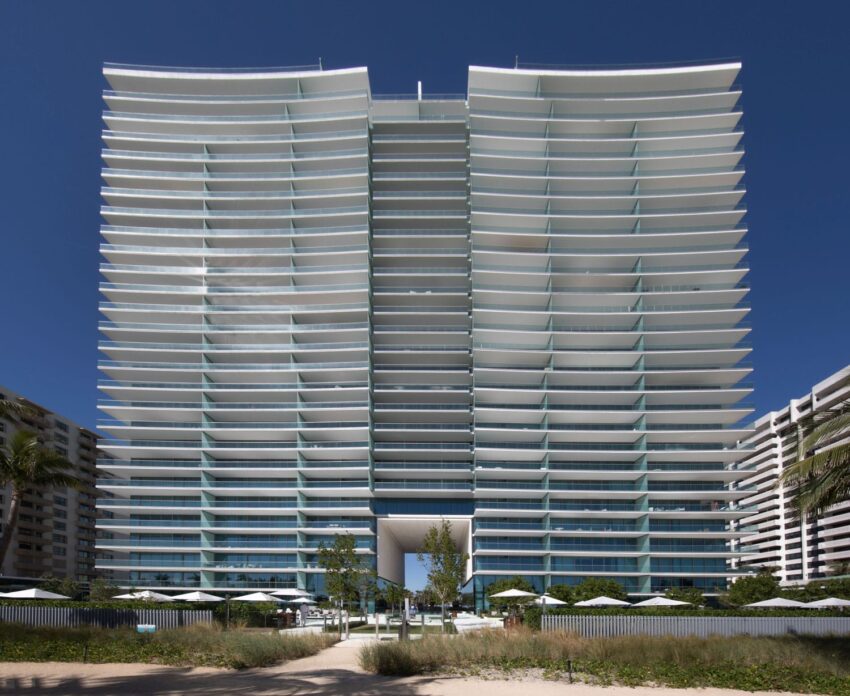Modern high-rise building with glass balconies under a clear blue sky, surrounded by palm trees and outdoor seating areas.