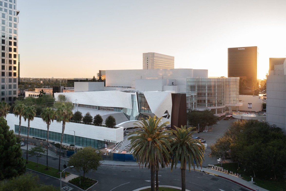 Aerial view of a modern architectural building surrounded by palm trees and cityscape at sunset.