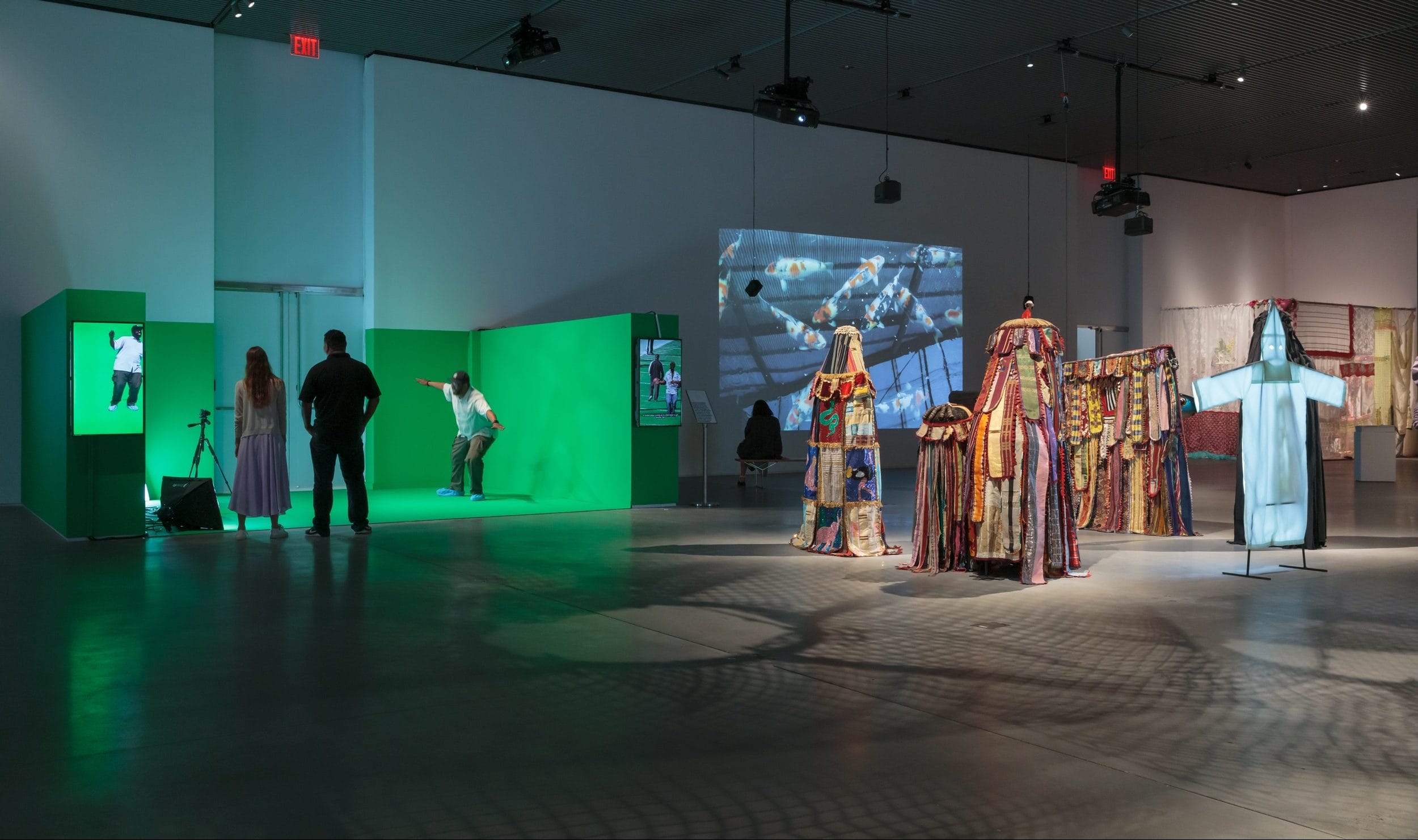 Art gallery with colorful costumes on display, people interacting with a green screen space, and projected video art on walls.