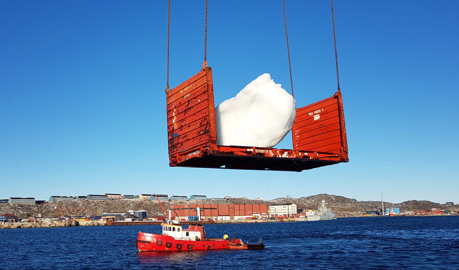 Crane lifting large iceberg piece from ocean with small red boat nearby and coastline buildings in the background.