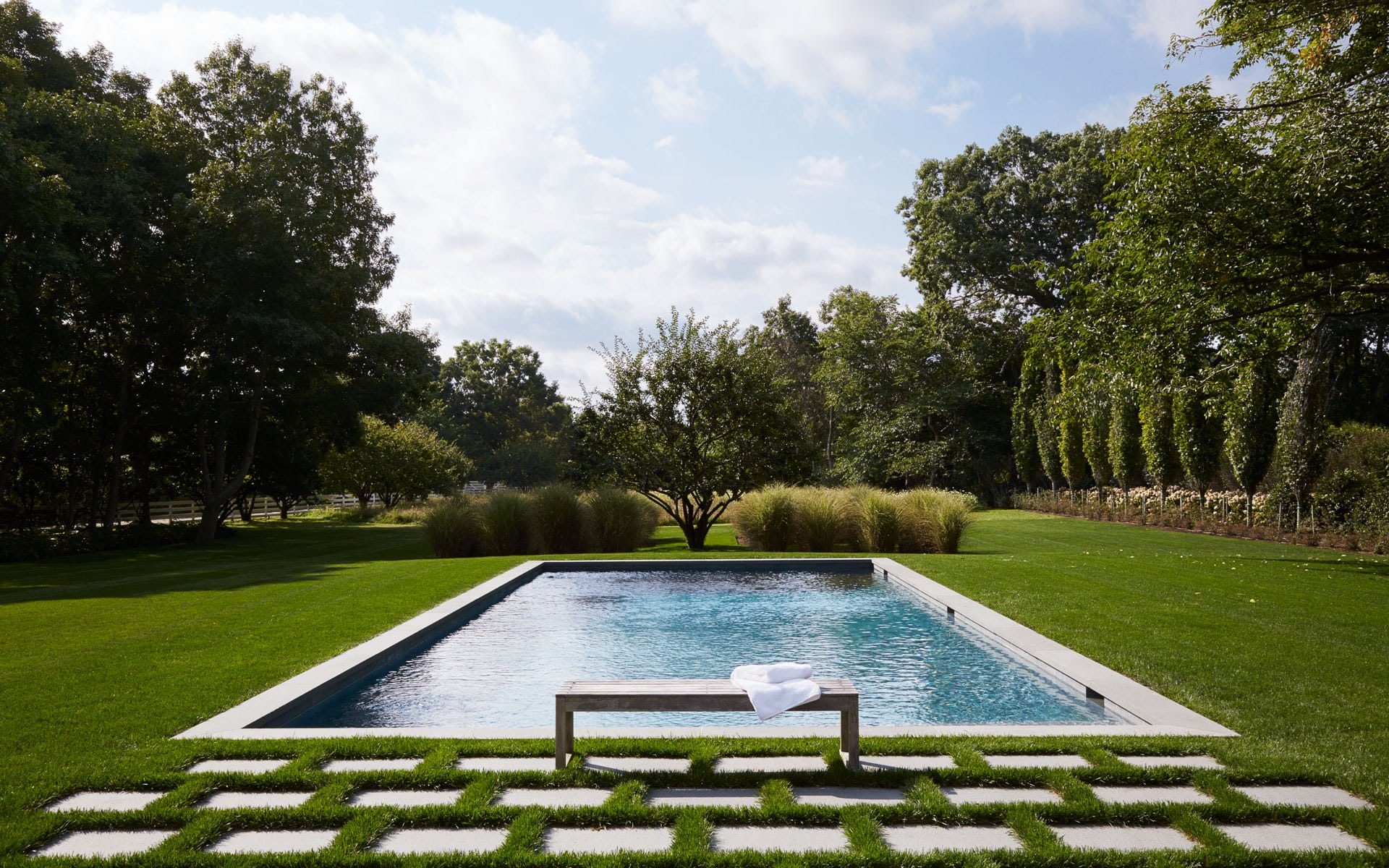 Rectangular swimming pool in a lush garden with a bench and trees in the background under a partly cloudy sky.