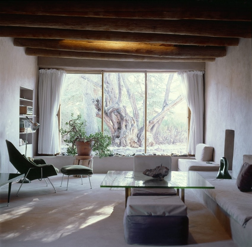 Sunlit living room with modern furniture, large window, wooden beams, white curtains, and desert view outside.