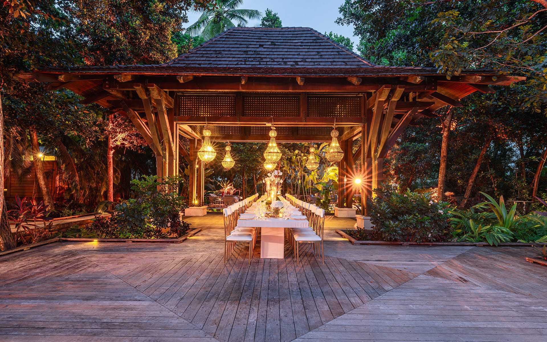 Outdoor pavilion with a long dining table set for an event, surrounded by trees and illuminated by hanging lanterns.