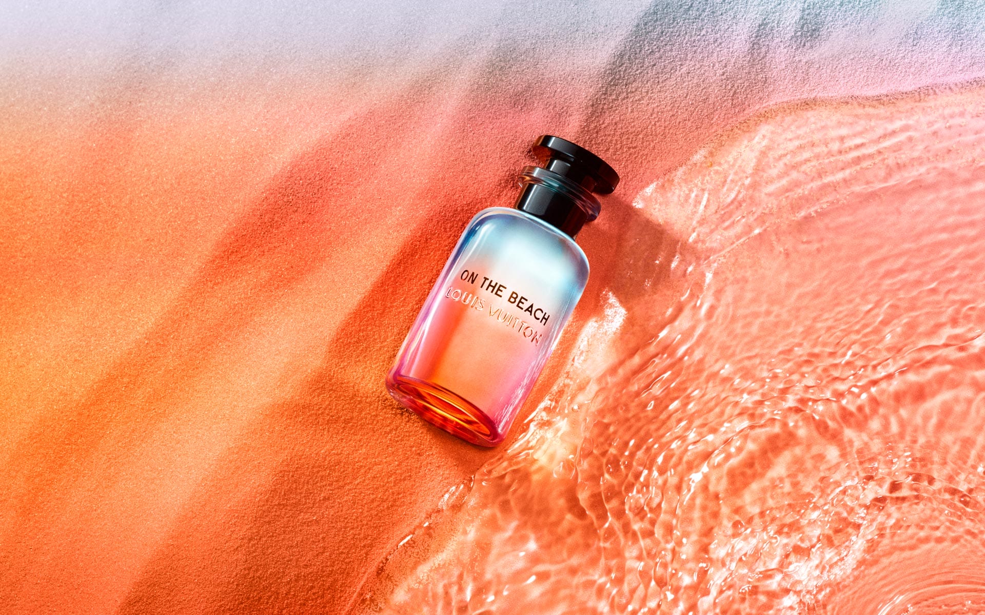 Perfume bottle labeled "On the Beach" by Louis Vuitton on sand with water reflecting sunlight.