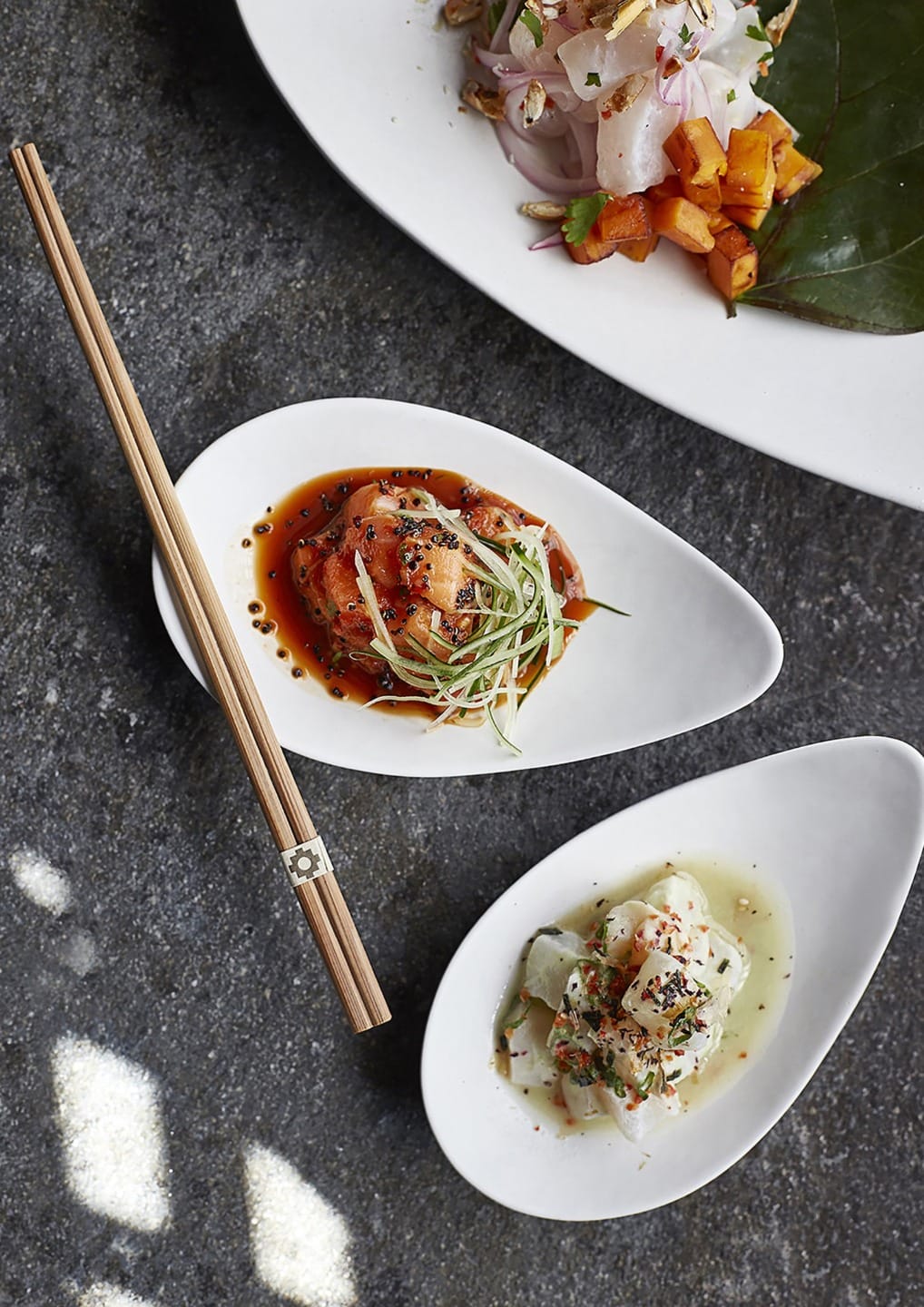 Two dishes of gourmet seafood appetizers with chopsticks on a dark surface, garnished with herbs and spices.
