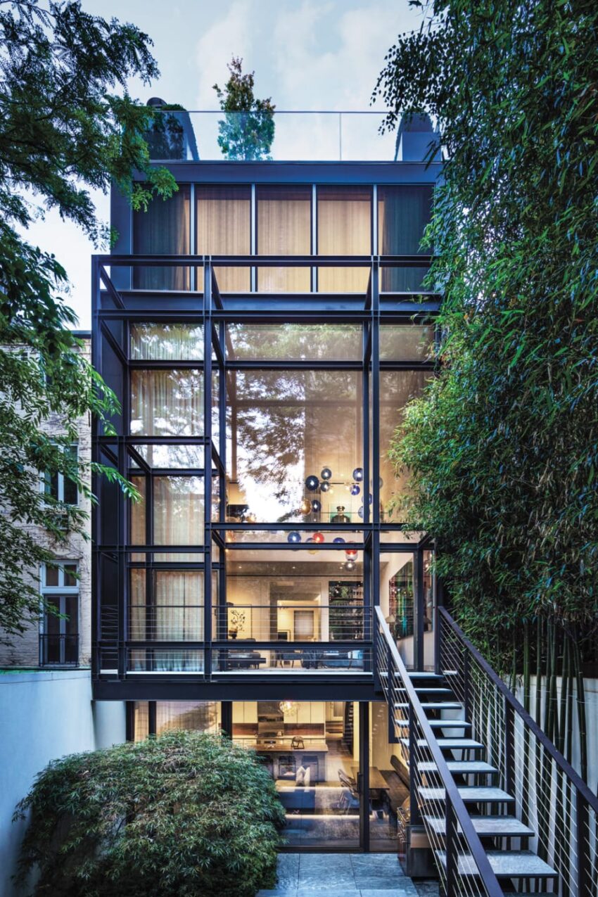 Modern multi-story glass house with external staircase, surrounded by lush greenery, showcasing interior design.