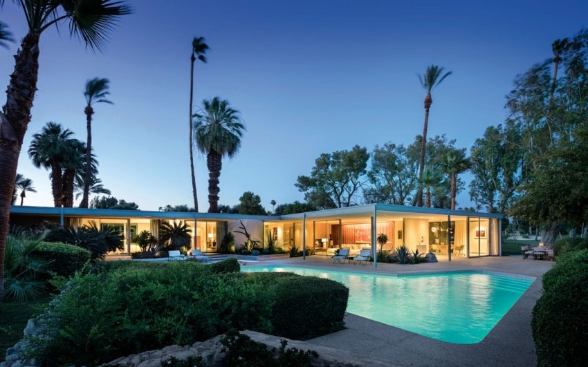 Modern house with large windows, surrounded by palm trees, and a glowing pool in the foreground at twilight.