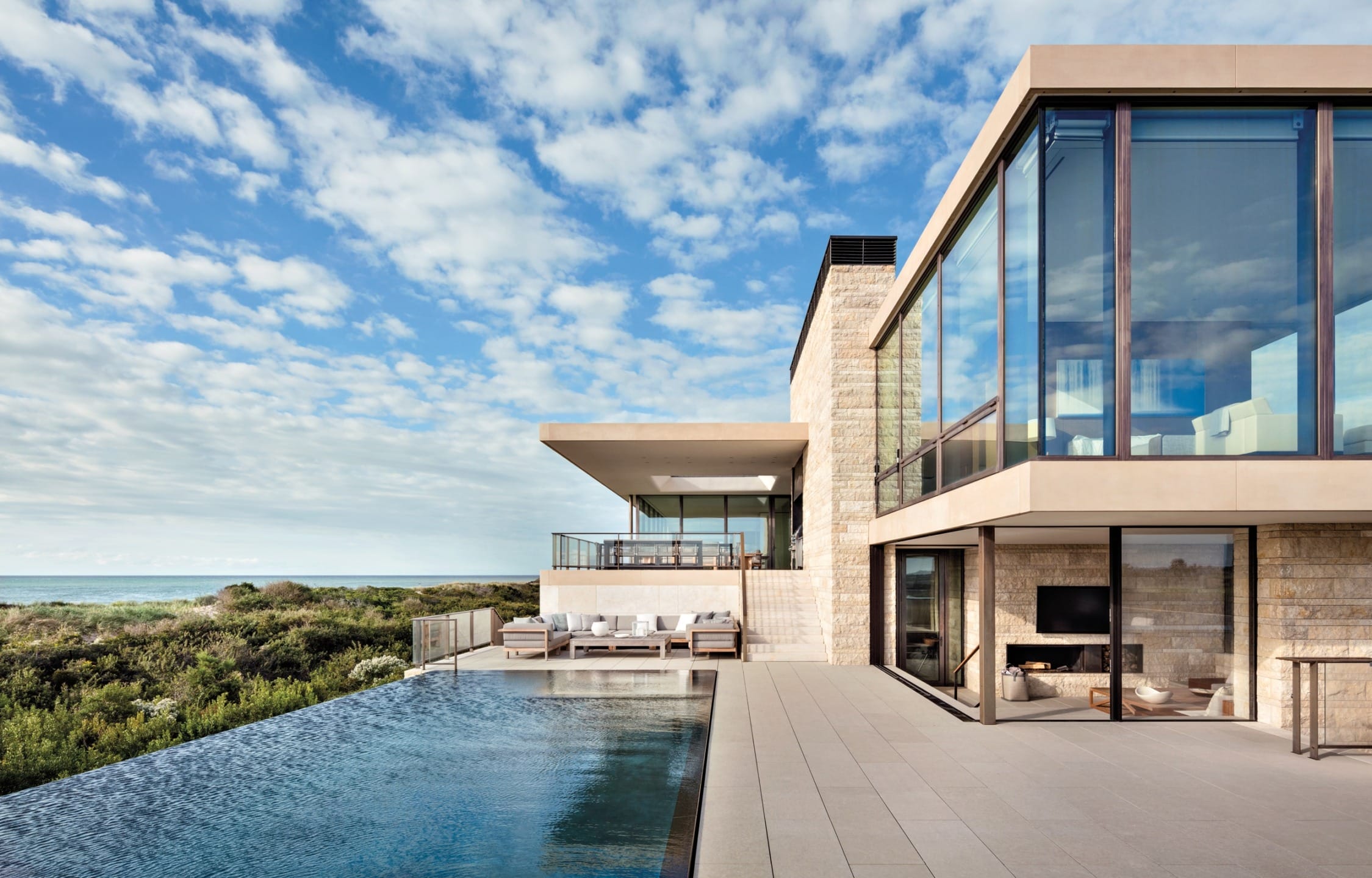 Modern seaside house with large windows, extended patio, and an infinity pool overlooking the ocean under a partly cloudy sky.