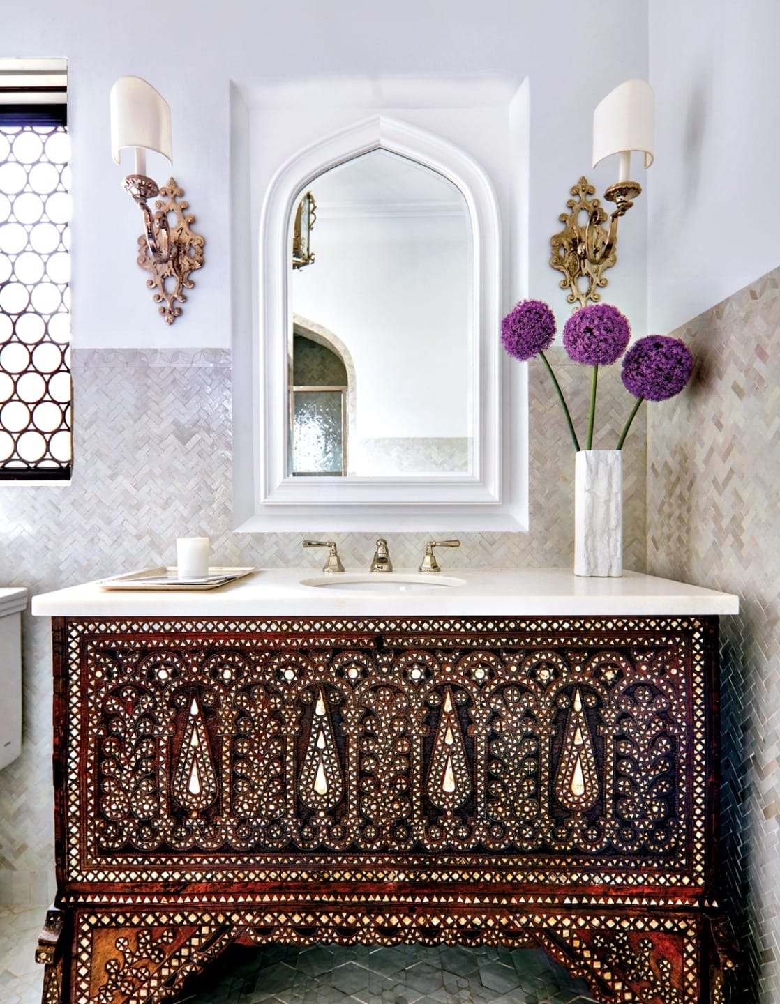 Ornate bathroom with detailed wooden vanity, vase of purple flowers, white mirror, and decorative tiled walls