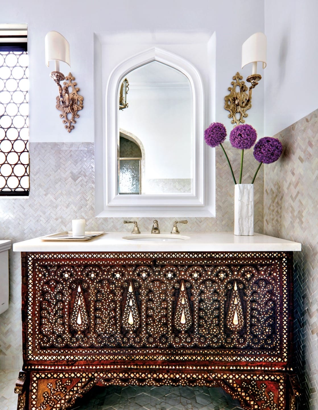 Ornate bathroom with detailed wooden vanity, vase of purple flowers, white mirror, and decorative tiled walls