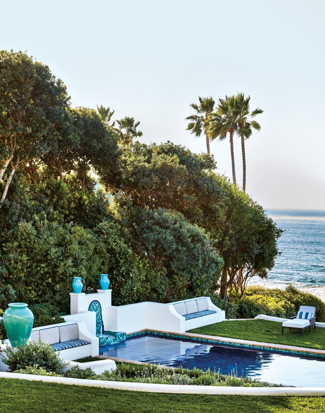 Luxurious outdoor pool with blue vases, white seating, ocean view, and palm trees surrounded by lush greenery.