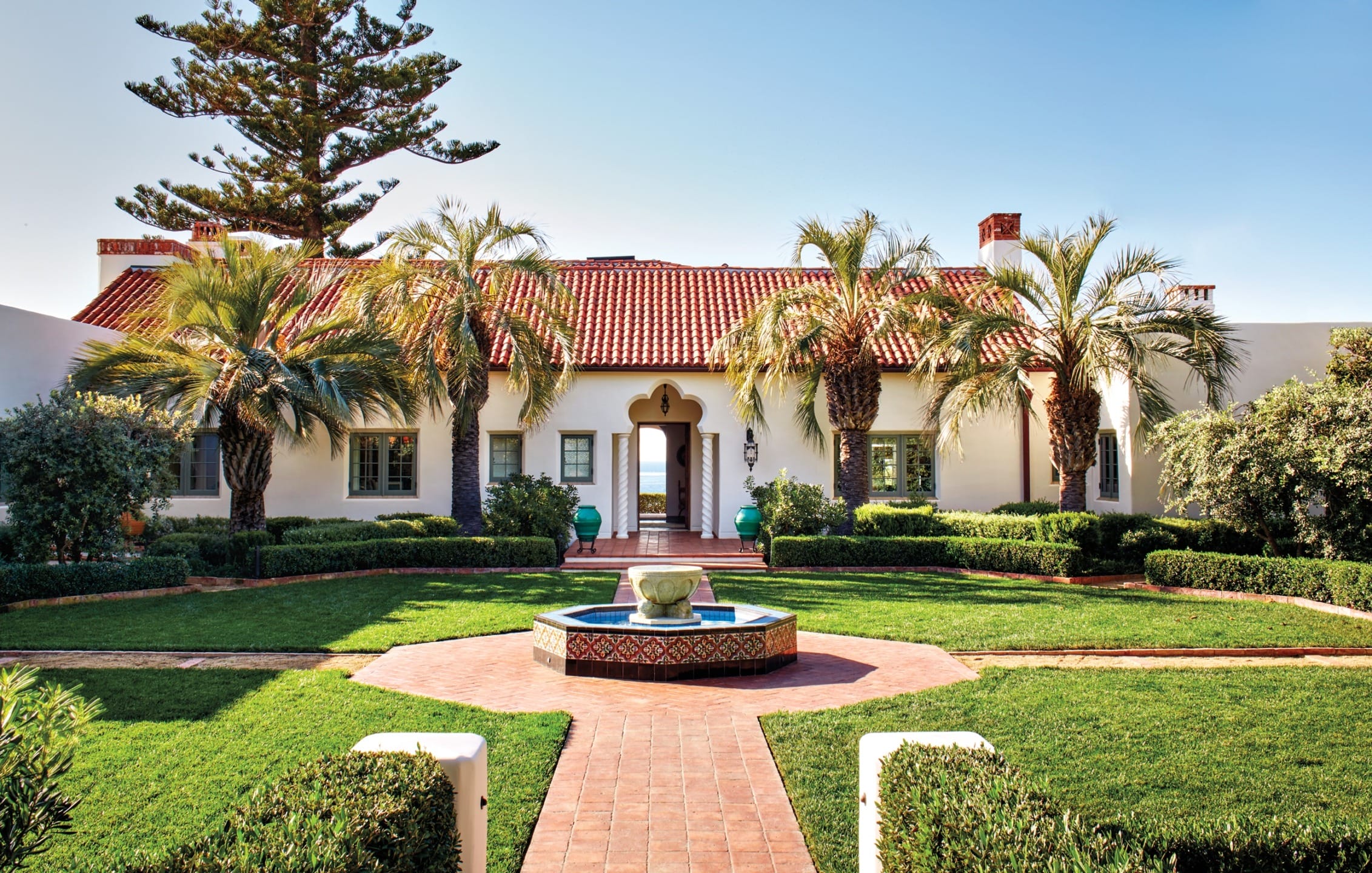 Spanish-style house with red tile roof, palm trees, and a decorative fountain in a landscaped garden setting.