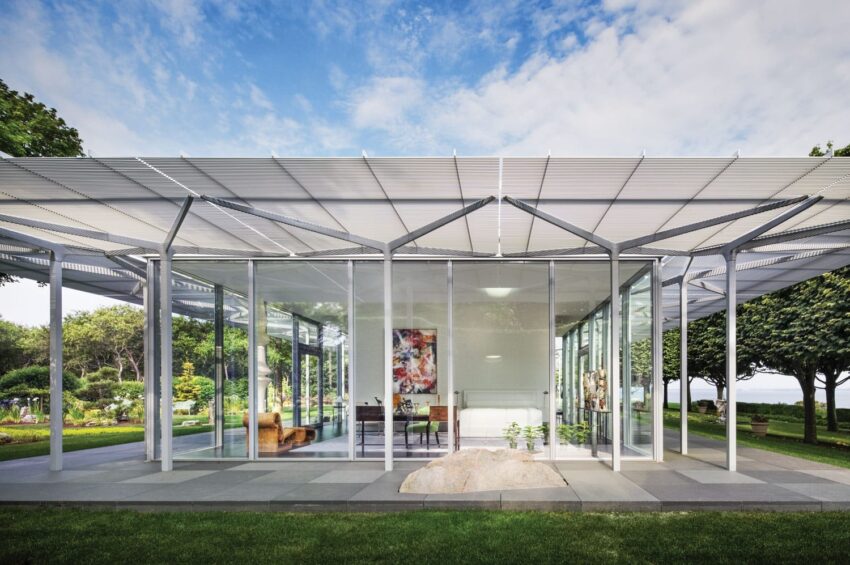 Modern glass pavilion with metal roof in a garden setting, featuring trees and a stone path in the foreground.