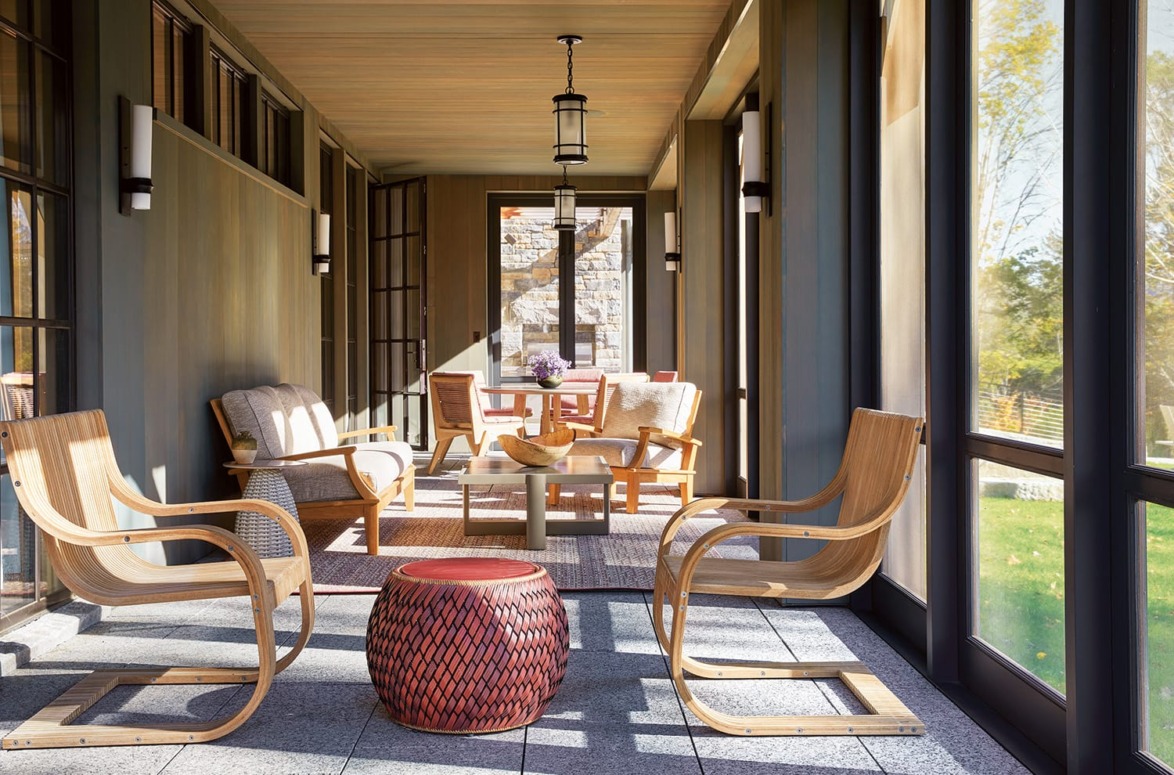 Spacious sunlit patio with wooden chairs, a red decorative table, and large windows overlooking greenery.