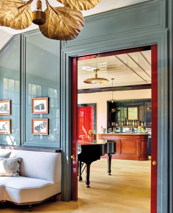 Elegant room with a gray sofa, gold leaf chandelier, and a grand piano visible through a red doorway leading to a bar area.