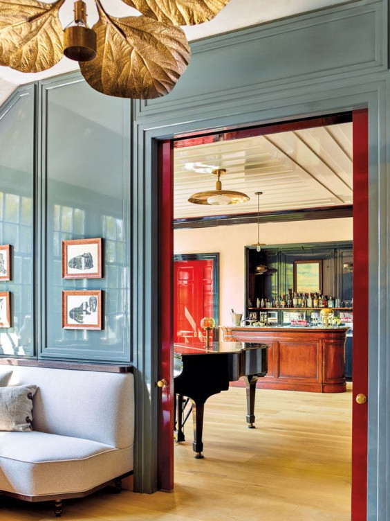 Elegant room with a gray sofa, gold leaf chandelier, and a grand piano visible through a red doorway leading to a bar area.