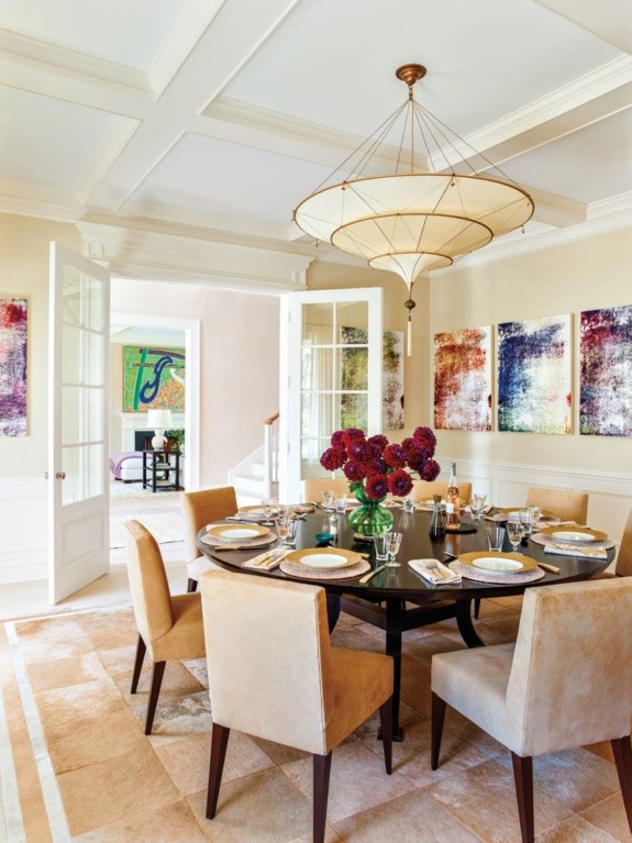 Elegant dining room with a round table, beige chairs, floral centerpiece, abstract paintings, and large chandelier.