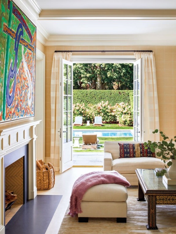 Elegant living room with open French doors leading to a garden, showcasing plush seating, a fireplace, and vibrant artwork.