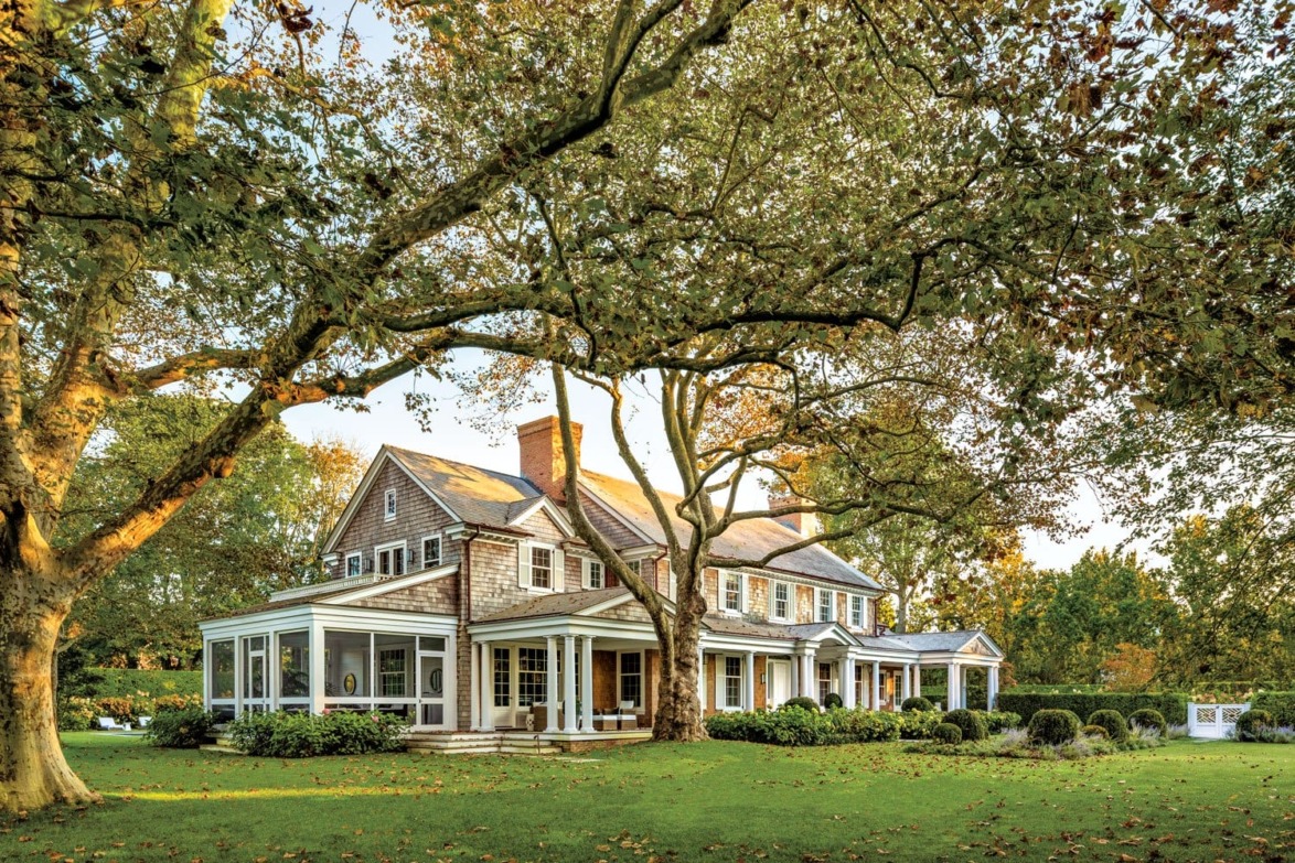 Charming two-story house with large trees in the front yard, surrounded by green grass, on a sunny day.