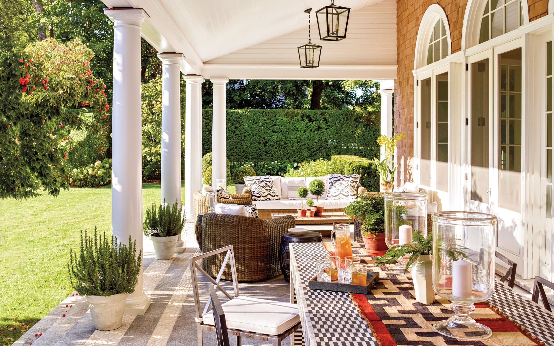 Sunny patio with wicker furniture, patterned cushions, dining table with candles, and a lush garden view in the background.
