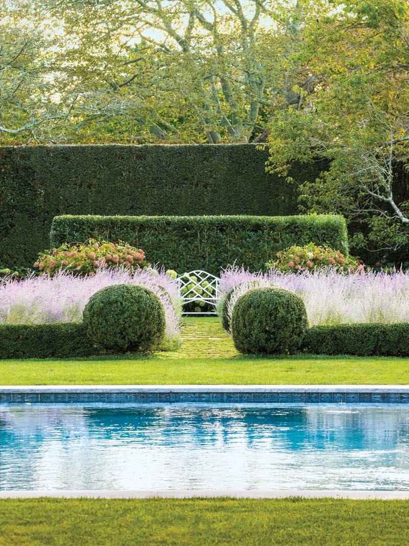 Serene garden with trimmed hedges, flowering plants, a white bench, and a clear blue pool in the foreground