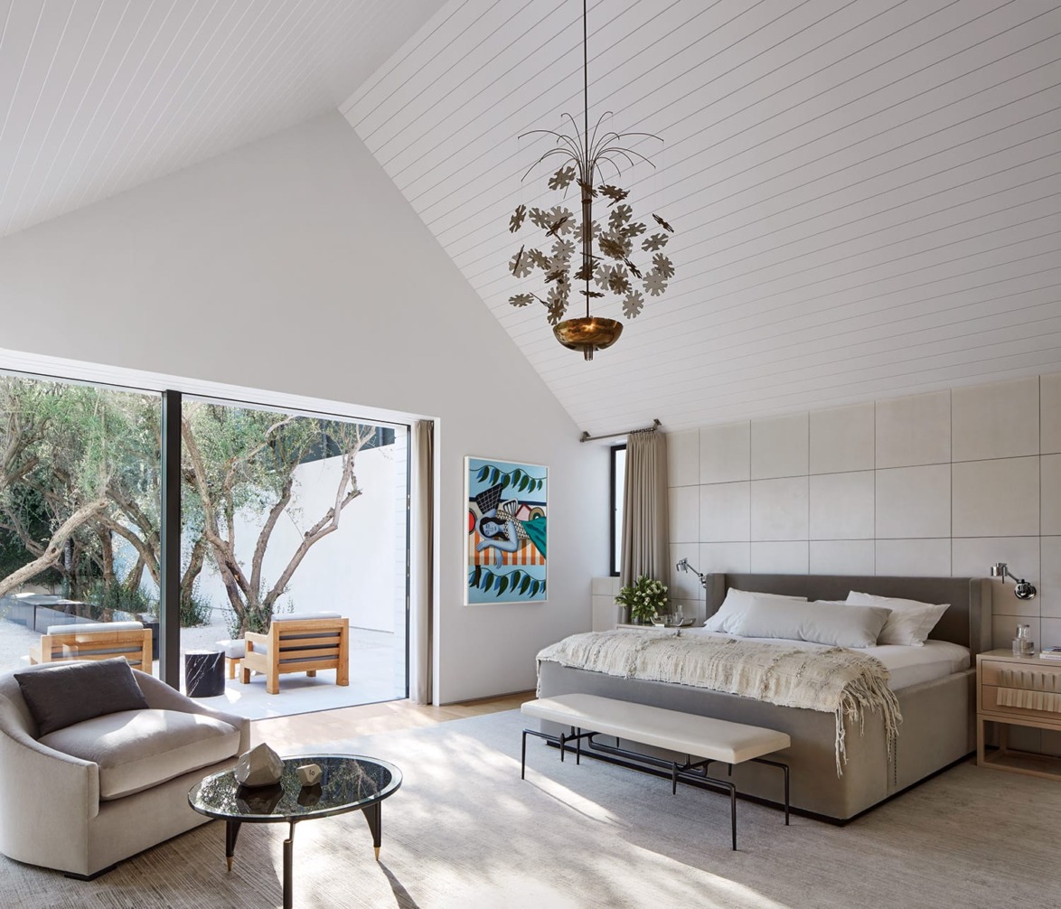 Spacious bedroom with modern decor, large bed, seating area, and sliding doors opening to a patio with wooden chairs and trees.