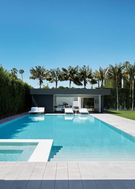 Luxurious backyard pool with sun loungers, surrounded by palm trees and modern pool house under a clear blue sky.