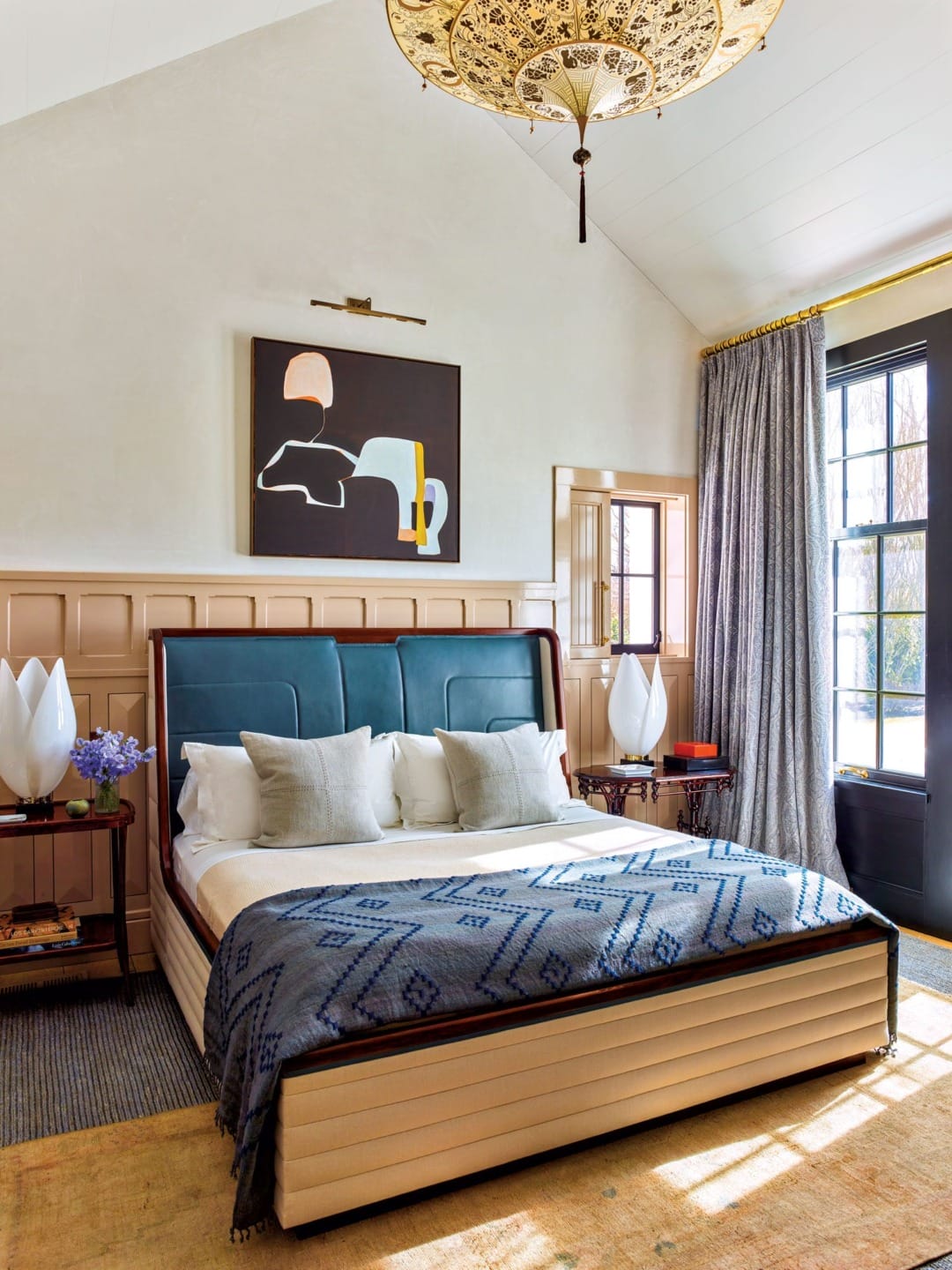 Elegant bedroom with modern artwork, teal headboard, patterned blanket, and large windows with gray curtains.