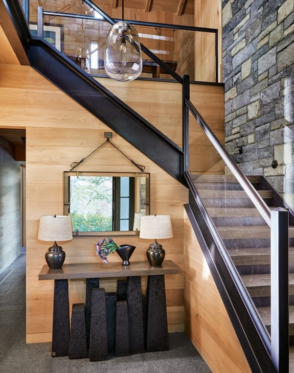 Modern foyer with wooden and stone walls, stylish black staircase, decorative console table with lamps, and a mirror.