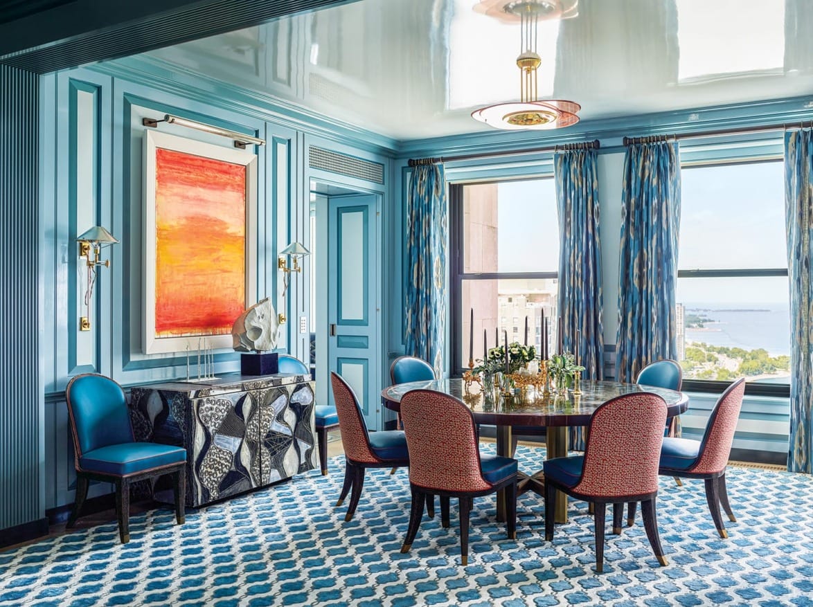 Elegant dining room with blue walls, abstract art, patterned carpet, and large windows offering a scenic view.