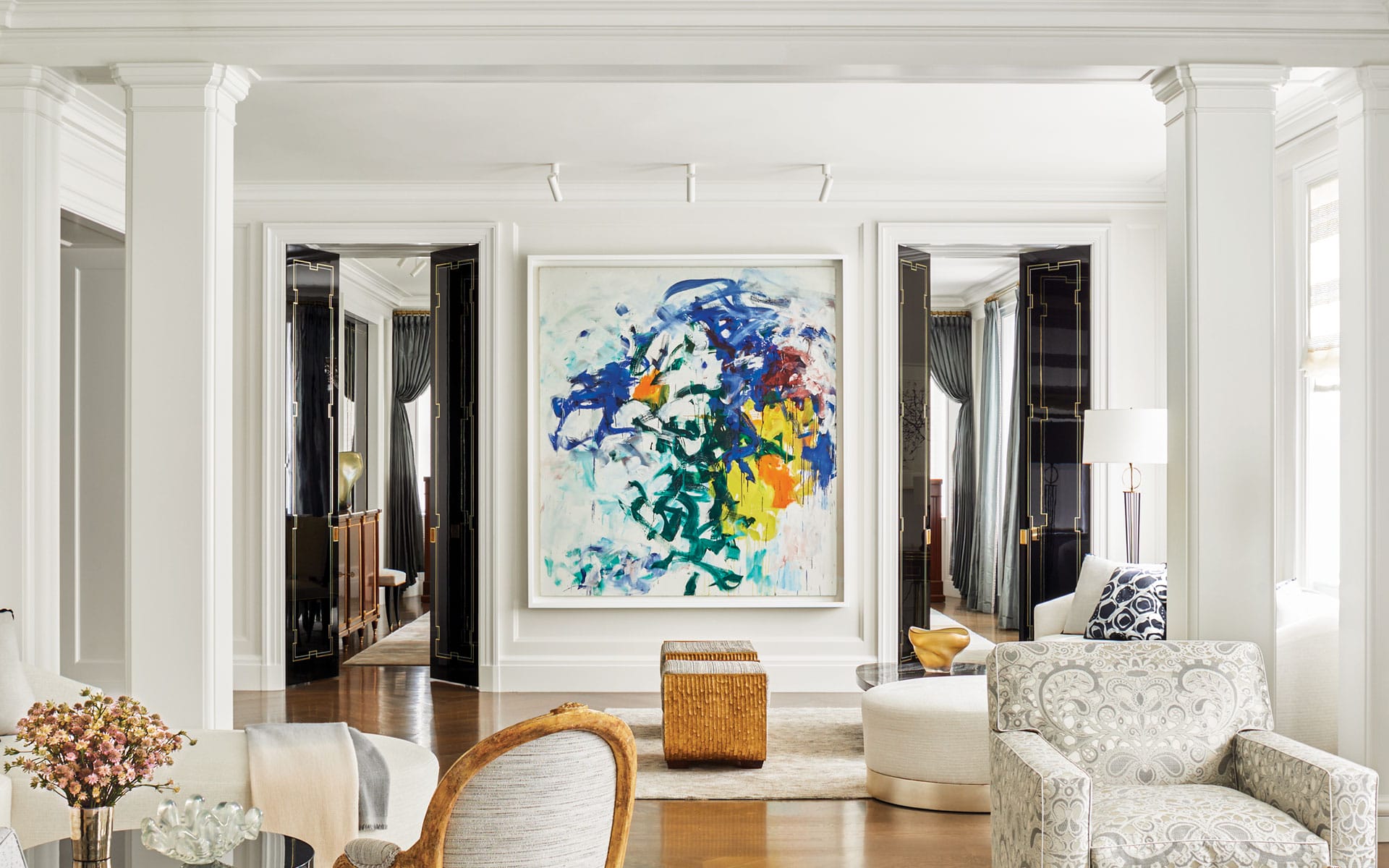 Elegant living room with abstract art, white walls, patterned chairs, wooden floors, and large windows with drapes.