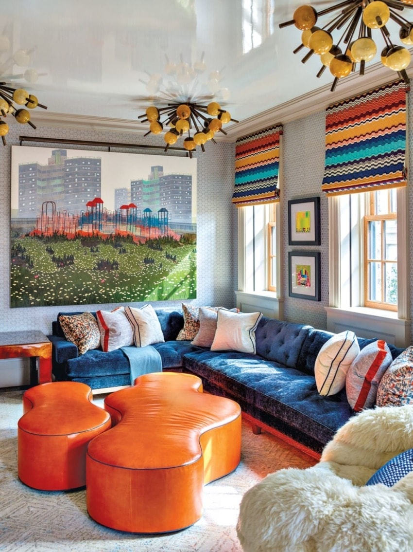 Colorful eclectic living room with modern artwork, blue sectional sofa, orange ottomans, patterned shades, and unique lighting.