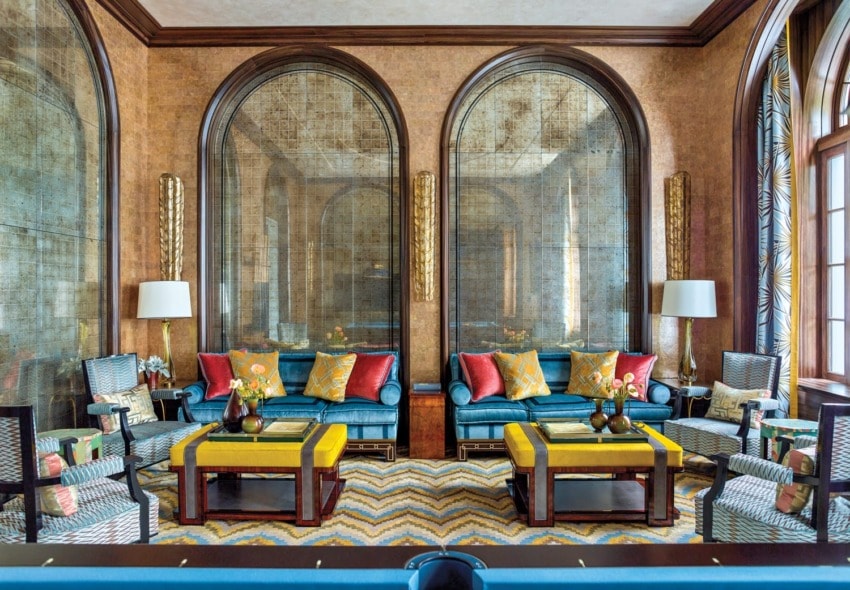 Elegant living room with blue sofas, yellow ottomans, patterned rug, large arched mirrors, and decorative lamps.