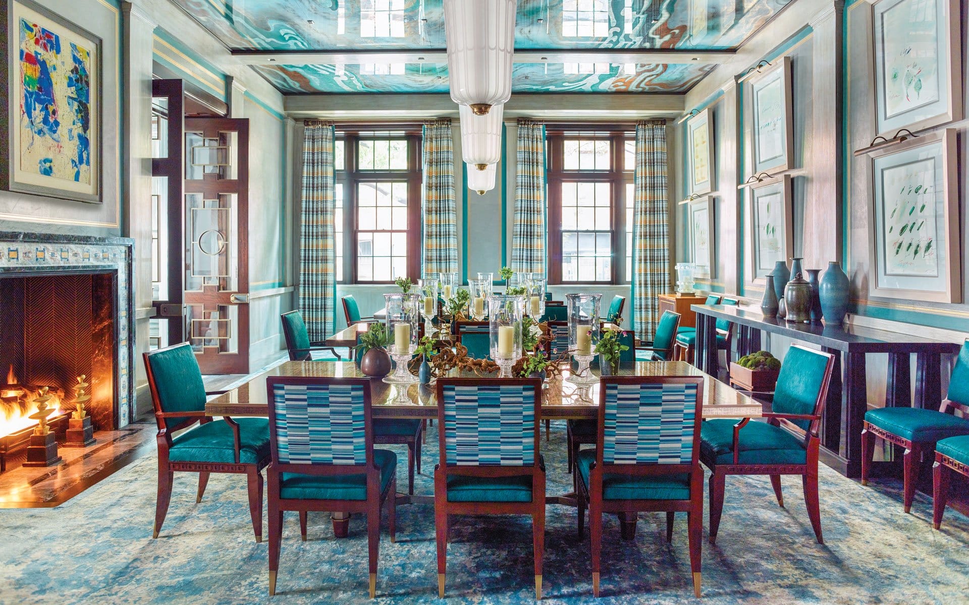 Elegant dining room with a long table, teal chairs, artwork, and a cozy fireplace, decorated in a modern style.