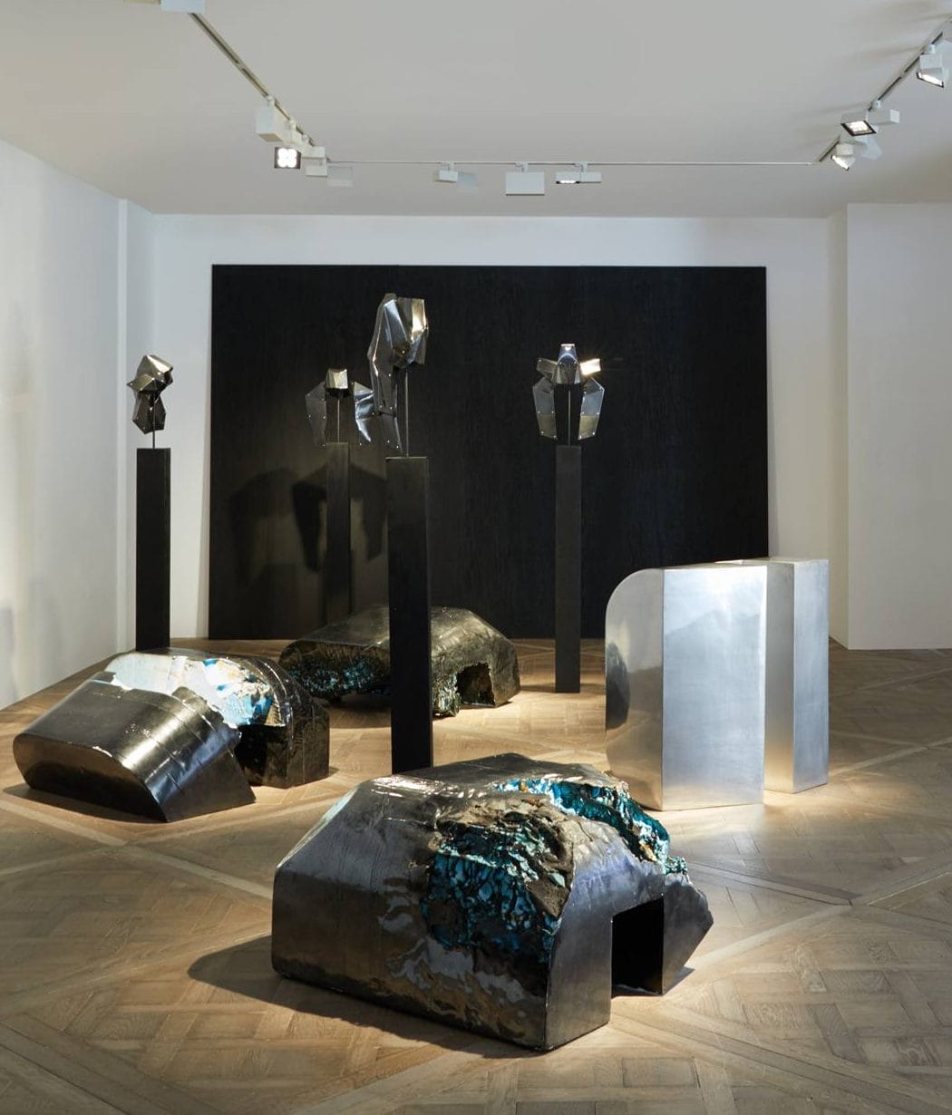 Contemporary art sculptures displayed in a gallery with abstract metallic forms on wooden flooring and dark backdrop.