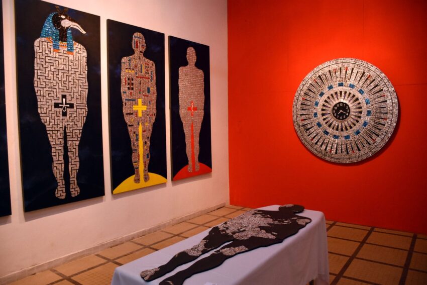 Art exhibit with four human-shaped maze paintings, a circular artwork on red wall, and a silhouette display on a white table.