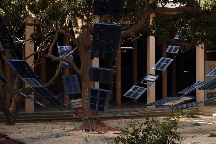 Art installation with blue panels hanging from a tree, surrounded by a wooden structure with sunlight filtering through.