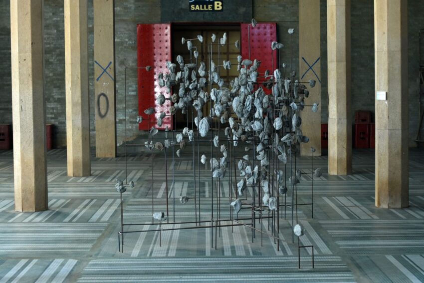 Art installation with scattered stones and metal rods in a spacious hall with tall pillars and patterned floor.