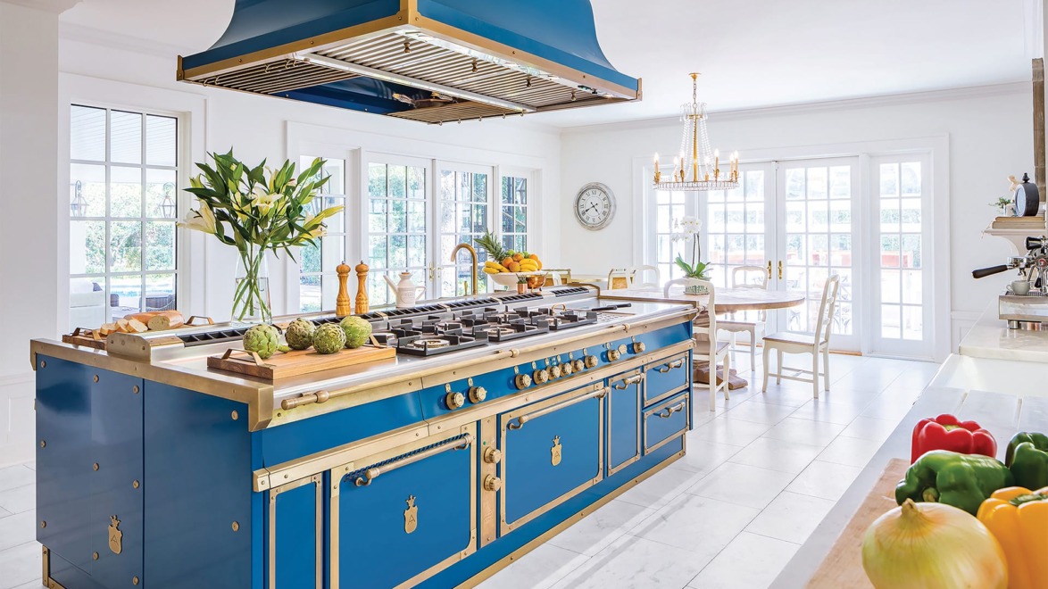 Luxury kitchen with blue stove island, chandeliers, fresh vegetables, and large windows for natural light.
