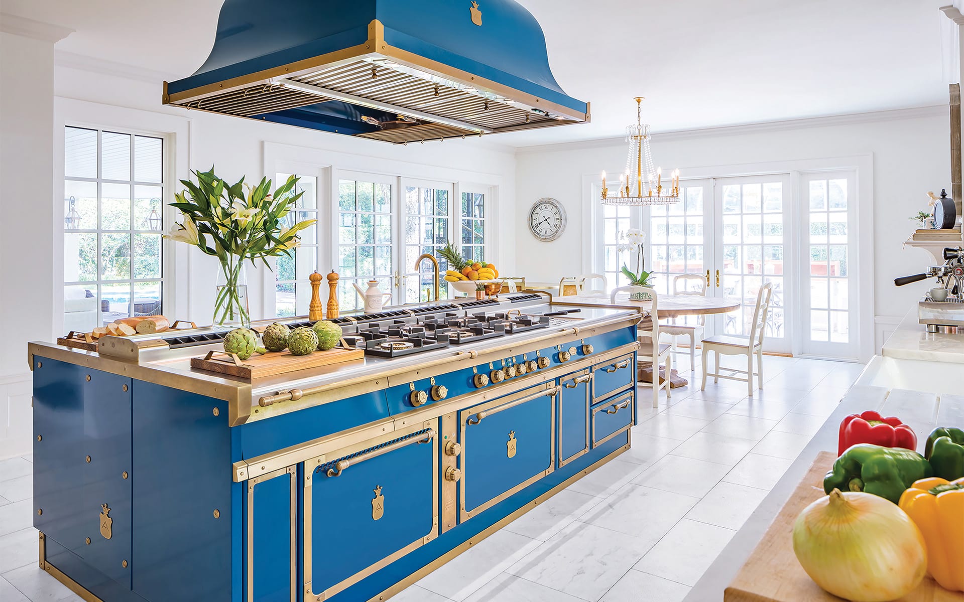 Luxury kitchen with blue stove island, chandeliers, fresh vegetables, and large windows for natural light.