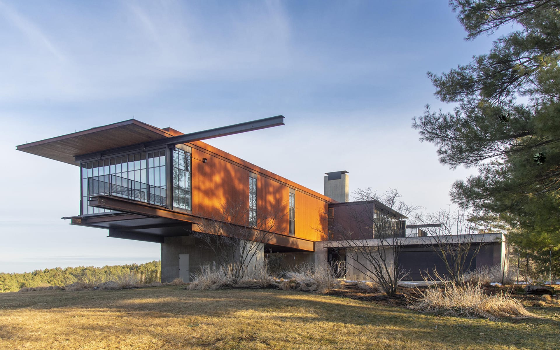 Modern architectural house with large glass windows and rust-colored exterior surrounded by trees and grass.