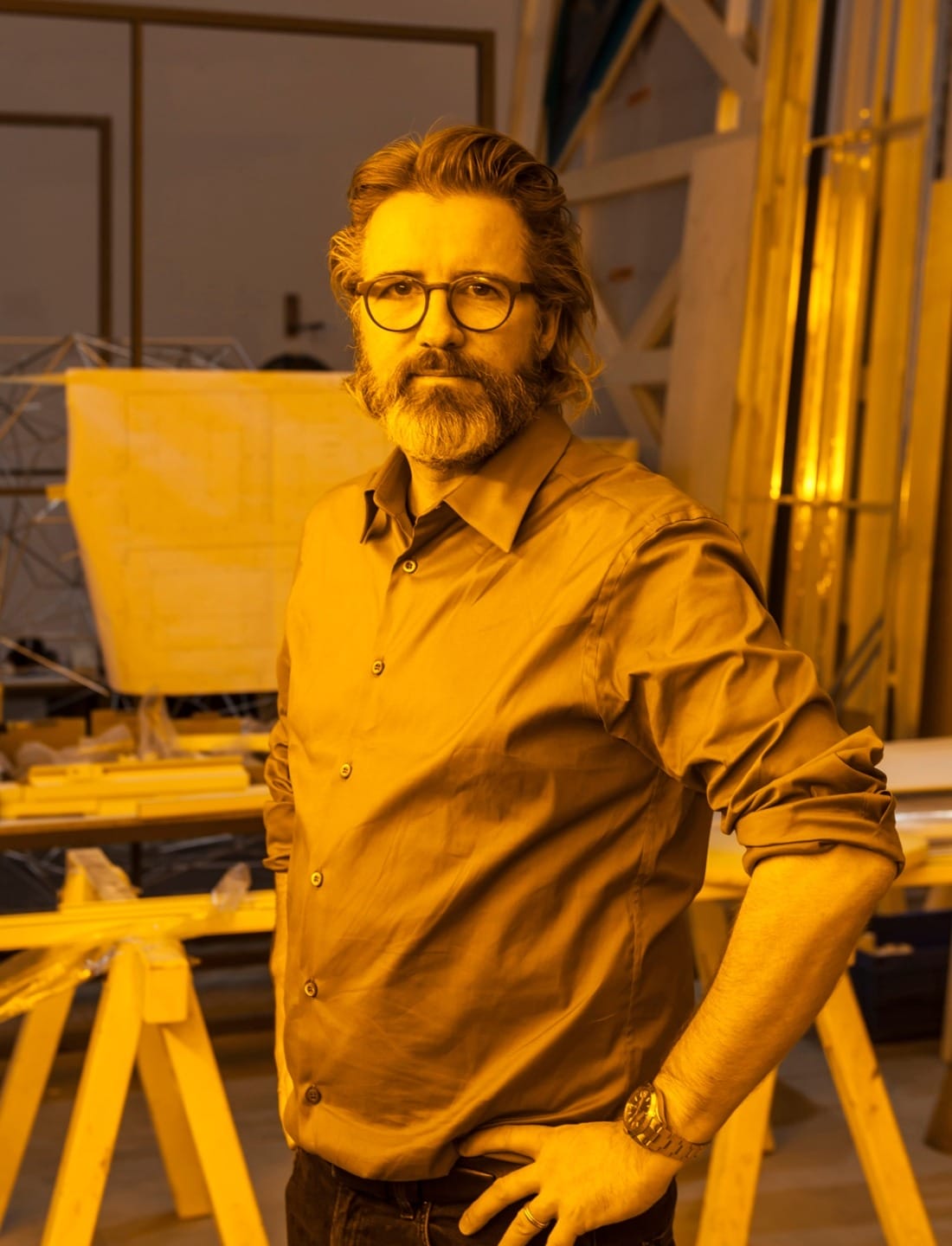 Man with glasses and beard in a workshop, wearing a shirt and posing with hand on hip, surrounded by wooden structures.