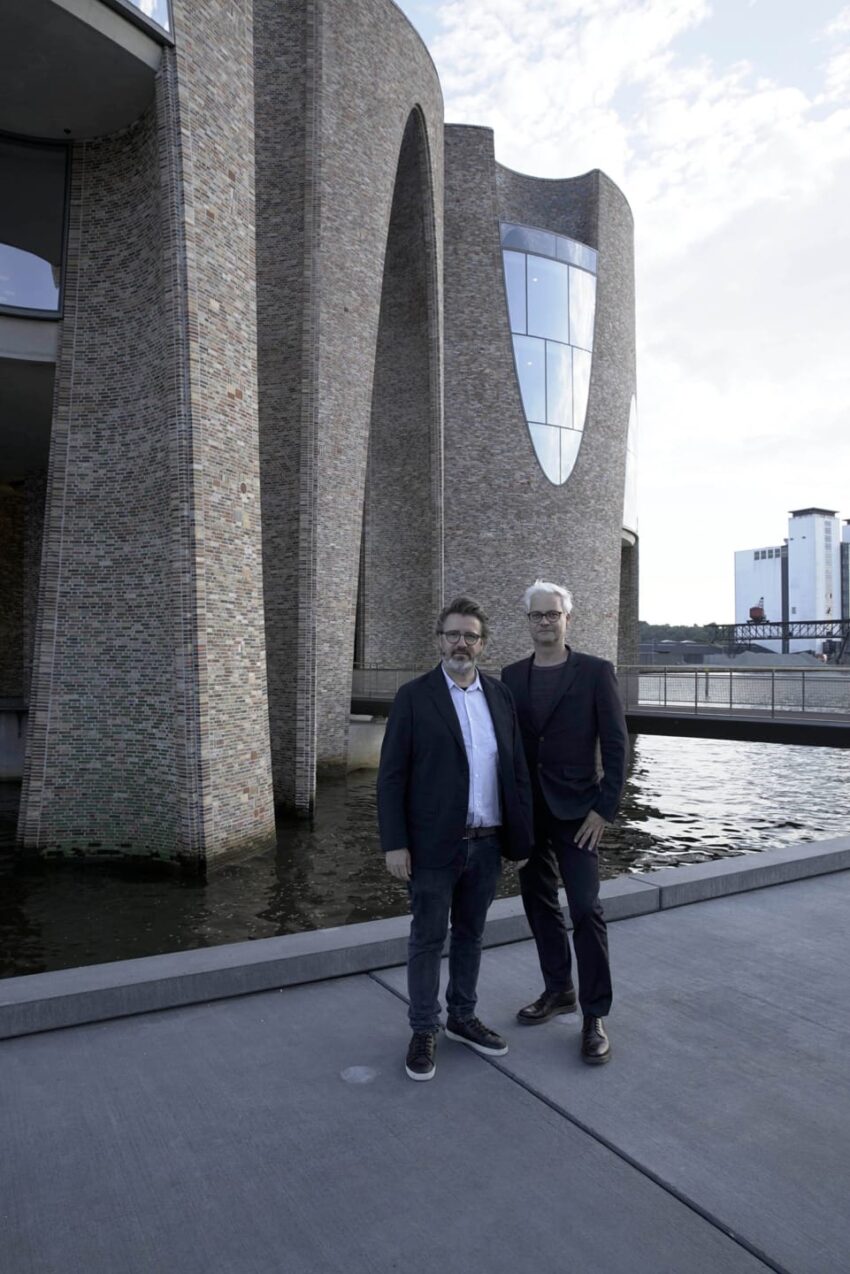 Two men standing in front of a modern brick building with large windows by the water.