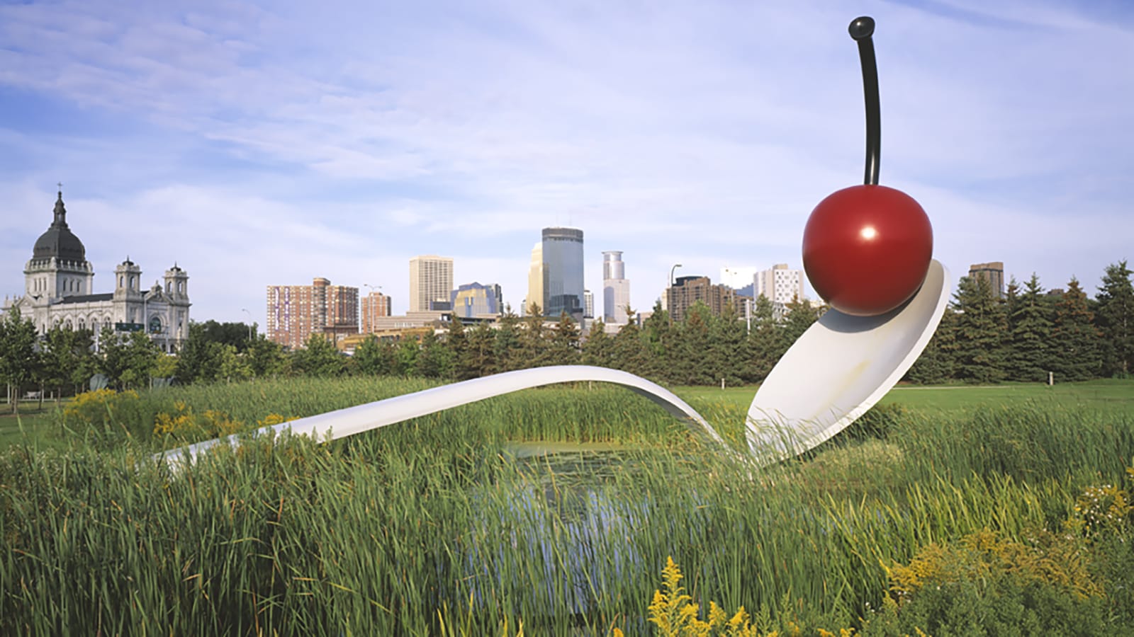 Spoonbridge and Cherry sculpture in a park with city skyline in the background.