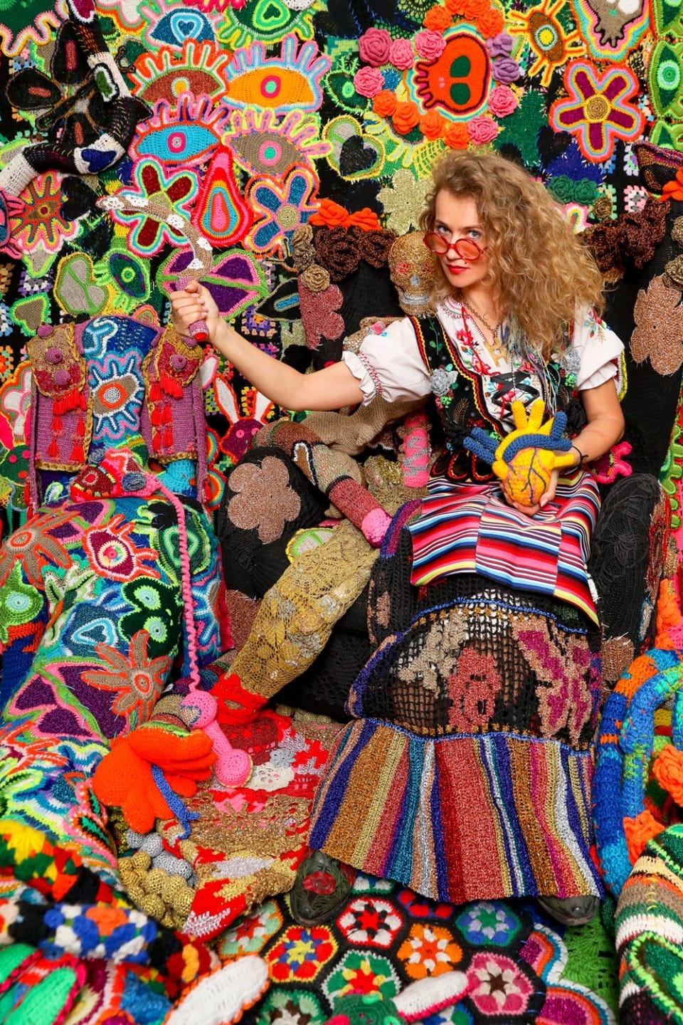 Person in vibrant, colorful crocheted outfit surrounded by intricate, multi-colored crochet patterns and designs.