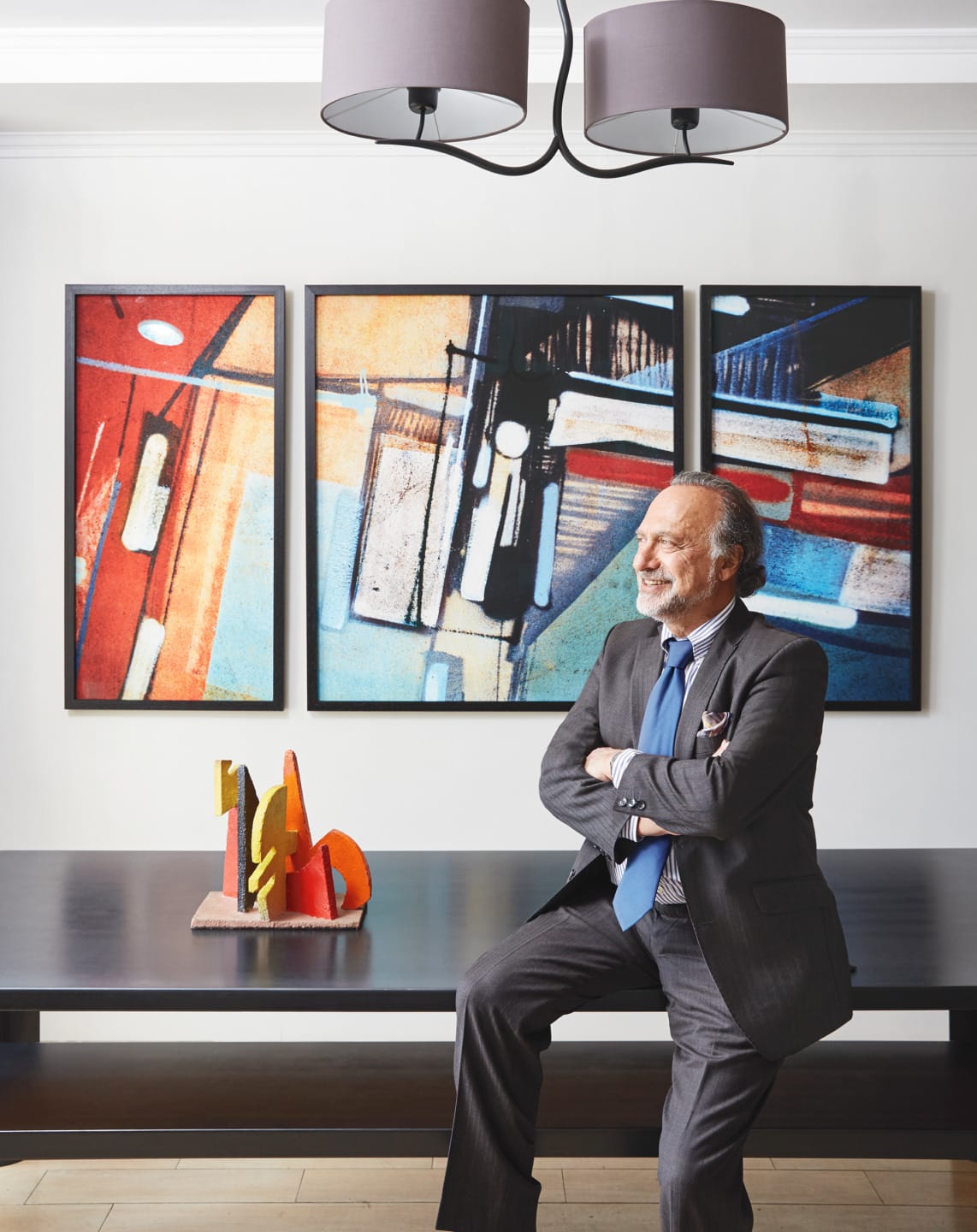 Man in suit sitting on table in modern art-filled room with abstract paintings and geometric sculpture.