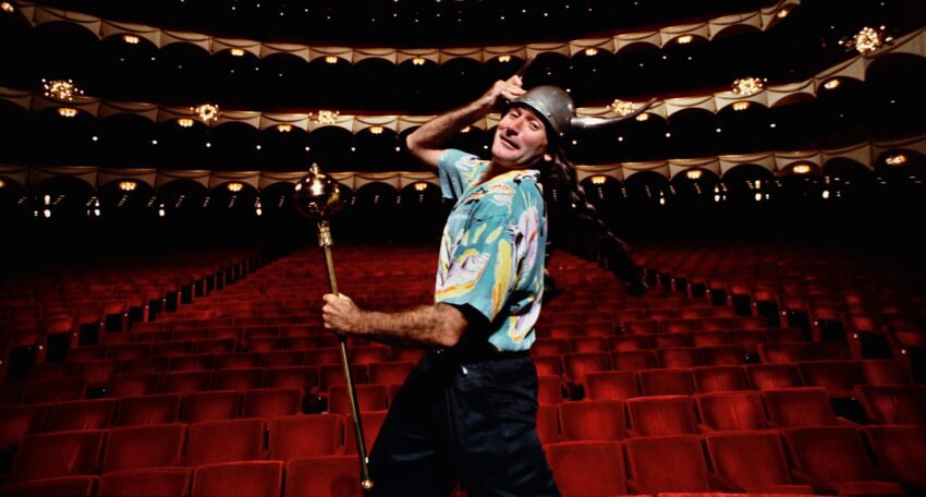 Entertainer in a colorful shirt and Viking helmet holding a staff, dancing in an empty theater with red seats and ornate decor.