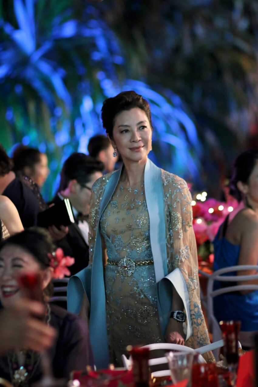 A person in an elegant dress standing at an outdoor event with colorful lighting and people seated around.
