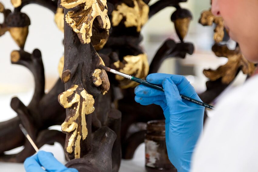 Person wearing blue gloves meticulously applying gold leaf to an ornate wooden sculpture.